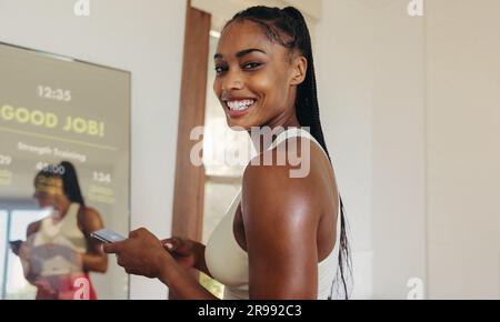 Une jeune femme heureuse et enthousiaste utilisant son smartphone et un miroir intelligent pour améliorer sa forme physique, en utilisant une technologie avancée pour l'atteindre Banque D'Images