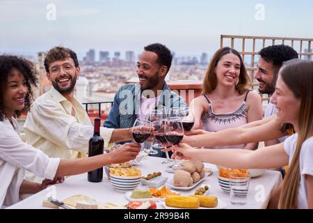Groupe multiethnique de personnes heureuses qui toaster avec du vin rouge sur un toit. Des amis qui célèbrent. Banque D'Images