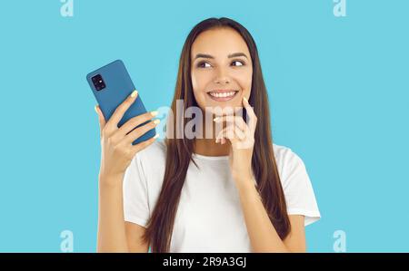 Une jeune fille amusante en t-shirt blanc tenant un smartphone et regardant de côté sur fond bleu. Banque D'Images