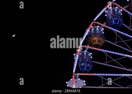 Vue sur une grande roue illuminée sur fond de ciel nocturne avec une lune Banque D'Images
