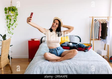 Bonne jolie femme prenant selfie avec le téléphone tout en préparant la valise de voyage Banque D'Images