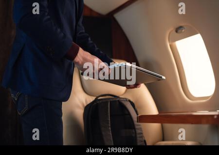 Un homme d'affaires gros plan, méconnu, sort un ordinateur portable moderne sous Windows et le place sur la table pour travailler à partir d'un avion privé de première classe Banque D'Images