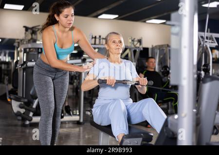 Une formatrice de forme physique féminine est engagée explique comment faire de l'exercice sur le simulateur à une femme âgée en salle de gym Banque D'Images