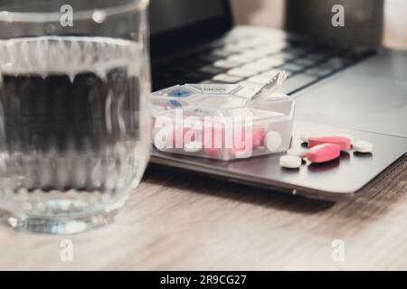 Organisatrice des prises de vue hebdomadaires avec un verre d'eau sur le lieu de travail et ordinateur portable gros plan de la boîte de pilules médicales avec des doses de comprimés pour la journée prendre des médicaments avec des médicaments blancs roses et des capsules. Médicaments compléments alimentaires, immunité et prévention des maladies huile de poisson gel Softgel, vitamine D et C minéraux oméga, fibres, acides aminés gras, probiotiques sédatifs. Les soins de santé et les maladies guérissent Banque D'Images