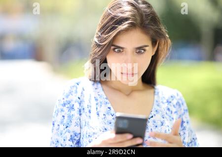 Vue de face portrait d'une femme perplexe regardant le téléphone des nouvelles surprenantes dans la rue Banque D'Images