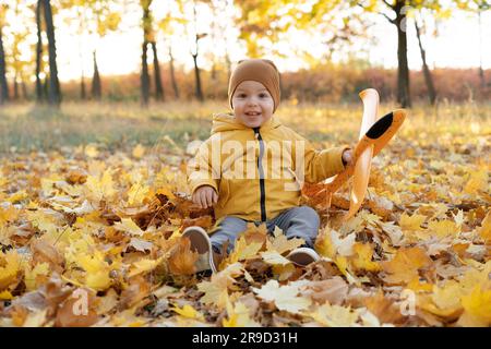 Un jeune garçon heureux s'assoit dans les feuilles mortes dans le parc d'automne. Détente en famille à l'extérieur en automne. Banque D'Images