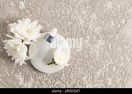 Flacon blanc mat avec compte-gouttes et bouchon en métal avec sérum ou produit cosmétique sur un plateau rond en marbre et des fleurs blanches. Vue de dessus. copier l'espace. Banque D'Images