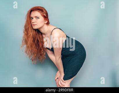 Femme à tête rouge fatiguée posant en studio sur fond gris. Cheveux longs, robe noire Banque D'Images