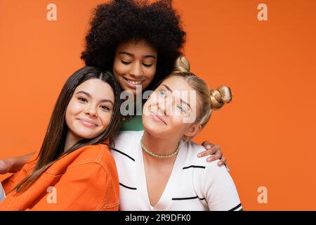 Modèle ado afro-américain souriant avec un maquillage brillant qui embrasse les filles à la mode et pose ensemble isolé sur l'orange, cool et confiant adolescent Banque D'Images