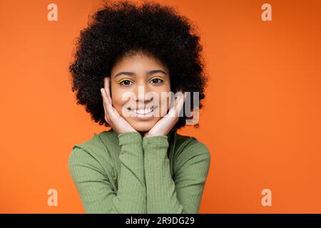 Portrait d'une jeune fille amérique africaine heureuse avec un maquillage audacieux portant une robe verte et touchant les joues tout en se tenant isolé sur l'orange, la culture de la jeunesse Banque D'Images