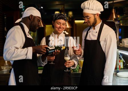 Un jeune chef afro-américain verse du champagne dans des flûtes tenues par ses deux collègues lors de la célébration de l'anniversaire de leur restaurant Banque D'Images