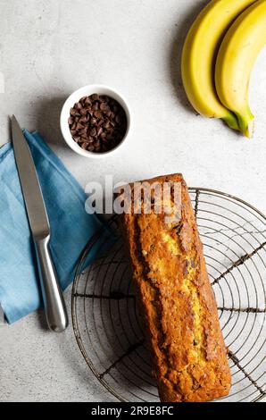 Un pain à la banane avec des pépites de chocolat, deux bananes, des pépites de chocolat dans un bol et une serviette bleue, sur fond gris. Banque D'Images