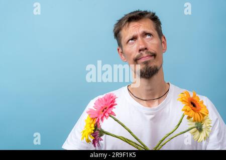 Portrait d'un adorable homme Caucasien réfléchi avec un bouquet de fleurs dans ses mains, avec un visage choqué réfléchi, regardant sceptique et exprimant d Banque D'Images