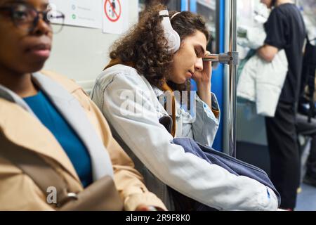 Vue latérale d'un adolescent dans un casque qui se serre contre la main courante tout en étant assis à côté d'une jeune femme dans le métro et en conduisant à l'école Banque D'Images
