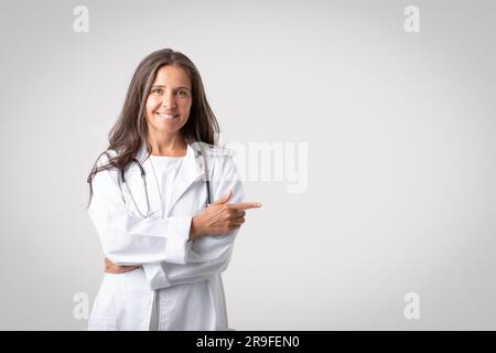 Gaie femme médecin senior en manteau blanc pointant le doigt sur l'espace libre isolé sur fond gris clair, bannière Banque D'Images