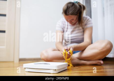 Femme en surpoids triste déprimé avec du ruban de mesure assis sur le sol près de l'échelle Banque D'Images