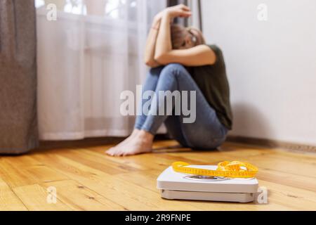 Femme triste et contrariée assise sur un sol en bois près de l'échelle blanche avec du ruban de mesure Banque D'Images