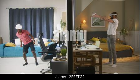 Les hommes multiraciaux, les voisins dans les micro-casques VR jouent à des jeux vidéo en réalité augmentée à l'aide de contrôleurs sans fil. Vue sur deux chambres ou appartements séparés par un mur. Concept de quartier et de style de vie. Banque D'Images