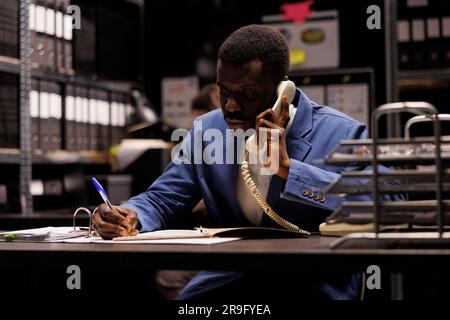 Un policier afro-américain parle avec un détective à distance à l'aide d'un téléphone fixe et parle d'indices confidentiels. Inspecteur privé analysant des preuves de scène de crime essayant de résoudre le cas dans la salle des arhives Banque D'Images