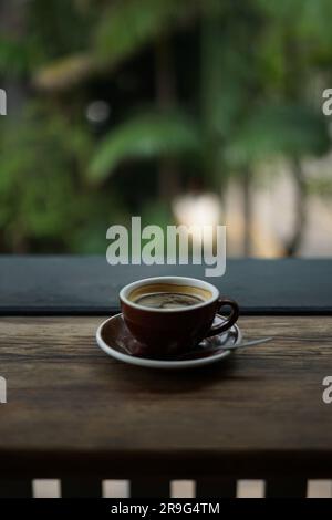 Une petite tasse brune sur une soucoupe remplie de café fraîchement préparé, placée sur une table en bois dans un cadre extérieur Banque D'Images