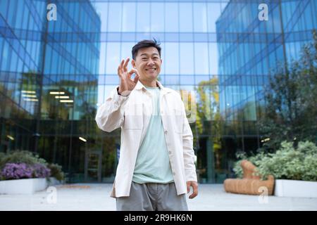 Homme asiatique heureux et réussi à l'extérieur du bâtiment de bureau souriant et regardant la caméra, montrant le signe ok avec les mains, programmeur satisfait de la réussite de la marche dans la ville urbaine moderne. Banque D'Images