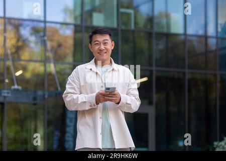 Portrait d'un homme asiatique heureux et réussi avec téléphone, homme souriant et regardant l'appareil photo, programmeur homme d'affaires utilisant l'application sur smartphone pour parcourir les pages Internet, marchant à l'extérieur du bâtiment de bureau. Banque D'Images