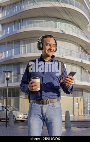 Jeune latin souriant avec un casque regardant son téléphone portable et tenant une tasse de papier avec un café dans la main dans une ville. Banque D'Images