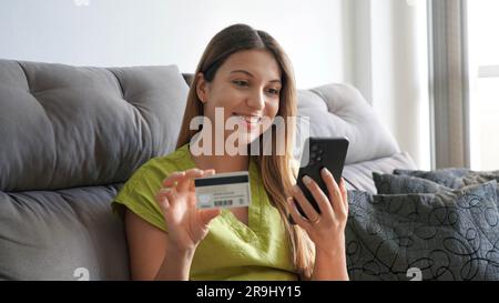 Bannière panoramique de fille gaie en utilisant un smartphone et une carte de crédit pour faire du shopping en ligne assis sur un canapé confortable à la maison. Banque D'Images