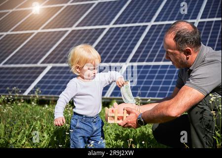 Son fils et son père économisent de l'argent et mettent dans la piggy banque. Père enseignant à son fils comment investir de l'argent à l'avenir sur fond de panneaux solaires. Concept d'économie d'argent, d'investissement dans les énergies renouvelables. Banque D'Images