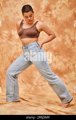 denim mode, génération z, jeune femme curvy avec tatouages posant avec la main dans la poche sur fond beige marbré, différentes formes, positivité du corps Banque D'Images