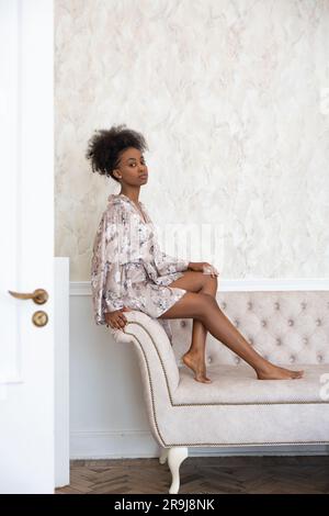 Jeune femme africaine en peignoir assis sur un canapé dans la chambre contre le fond de l'intérieur de la maison. Banque D'Images