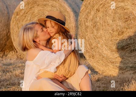 Maman embrasse sa fille bien-aimée assise dans un champ près d'une botte de foin. Mère et fille aimantes. Loisirs de plein air en famille. Banque D'Images