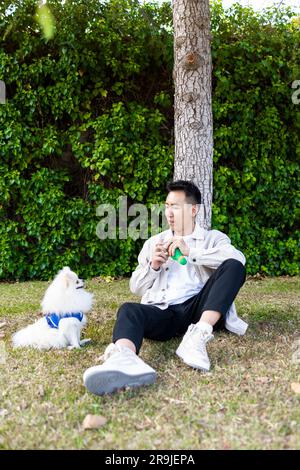 Tout le corps de positif asiatique propriétaire dans des vêtements décontractés assis sur le jeu avec le chien de Pomeranian dans le parc vert Banque D'Images