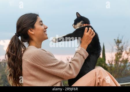 Vue latérale d'une femme charmante souriant et regardant un adorable chat blanc et noir adopté tout en se liant sur le toit Banque D'Images
