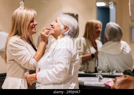 Vue latérale d'une femme mûre et gaie appliquant un rouge à lèvres sur les lèvres d'une femme âgée souriante près du miroir dans la salle de bains Banque D'Images