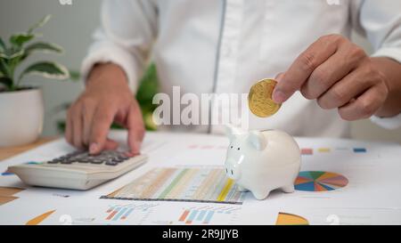 Homme mettant des pièces de monnaie dans la banque de porc et utilisant le calculateur d'impôt sur le revenu personnel pour payer des impôts. Recherche financière. Rapport. Calcul de la déclaration de revenus. Banque D'Images