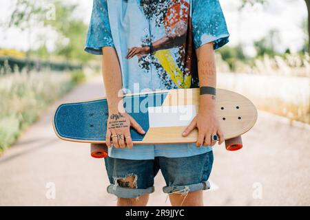 Jeune femme courte et non reconnaissable avec un ensemble décontracté de tomboy tenant un skateboard debout dans la rue et sur un arrière-plan flou Banque D'Images