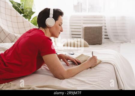 Vue latérale d'une femme mûre concentrée en robe rouge, posée sur un lit moelleux et écrivant des informations dans un ordinateur portable tout en écoutant de la musique dans un casque Banque D'Images
