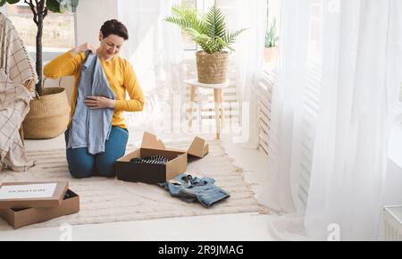 Corps complet de vêtements pliants d'âge moyen souriants tout en l'emballant dans une boîte en carton pour le don dans le salon léger Banque D'Images