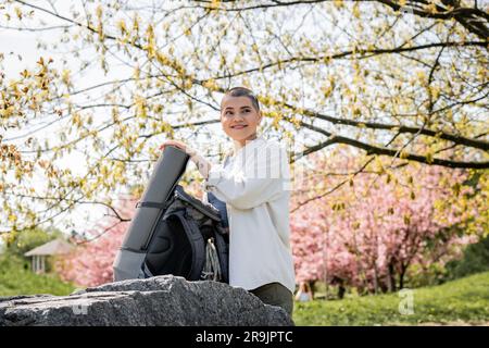 Heureuse jeune femme petite cheveux et tatouée voyageur dans des vêtements décontractés regardant loin en se tenant près de sac à dos avec l'équipement de voyage sur la pierre avec Banque D'Images