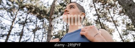 Vue à angle bas de la femme à poil court et tatouée randonneur tenant un sac à dos et regardant loin en se tenant dans une forêt d'été floue, Wanderlust vibes co Banque D'Images