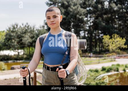 Portrait de jeune femme touristique aux cheveux courts et tatouée avec sac à dos regardant l'appareil photo tout en tenant des bâtons de randonnée avec la nature à l'arrière-plan, REC Banque D'Images