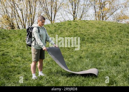 Souriant jeune femme court aux cheveux touriste dans des vêtements décontractés avec sac à dos mettant le tapis de fitness sur l'herbe avec la colline et la nature à l'arrière-plan, la connexion Banque D'Images