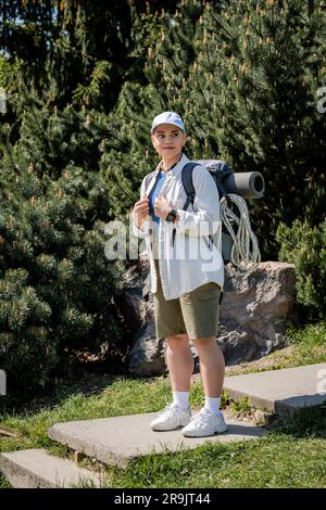 Jeune femme positive en casquette de baseball et vêtements décontractés tenant un sac à dos et regardant loin tout en se tenant debout avec la nature à l'arrière-plan, curieux Banque D'Images