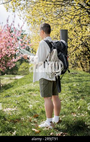 Jeune petite fille aux cheveux et tatouée touriste avec sac à dos, tapis de fitness et équipement de voyage tout en tenant la carte et debout avec la nature à backgroun Banque D'Images