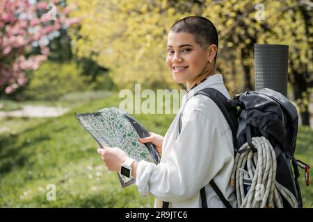 Souriant court aux cheveux et tatoué femme touriste dans des vêtements décontractés avec sac à dos et de l'équipement de voyage tenant la carte et regardant l'appareil photo avec la nature à Banque D'Images