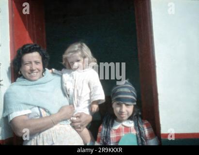 Venezuela, fin 1950s: Mère debout dans la porte de la maison tenant la fille blonde aux cheveux dans la chemise de nuit blanche, fille plus âgée debout portant une robe de praid Banque D'Images