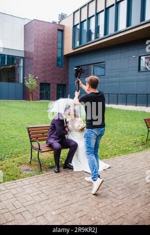 Un vidéaste professionnel de mariage photographie un film avec les jeunes mariés sur un banc sous un parapluie Banque D'Images