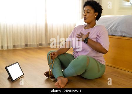 Femme afro-américaine focalisée de taille plus pratiquant la méditation de yoga à l'aide de tablette, espace de copie à la maison Banque D'Images