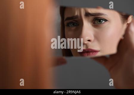 Jeune femme se regardant dans un éclat de miroir cassé sur fond gris, gros plan Banque D'Images